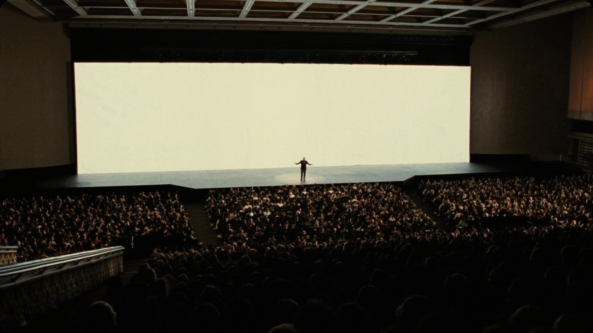 A speaker alone on a wide stage addressing a full auditorium